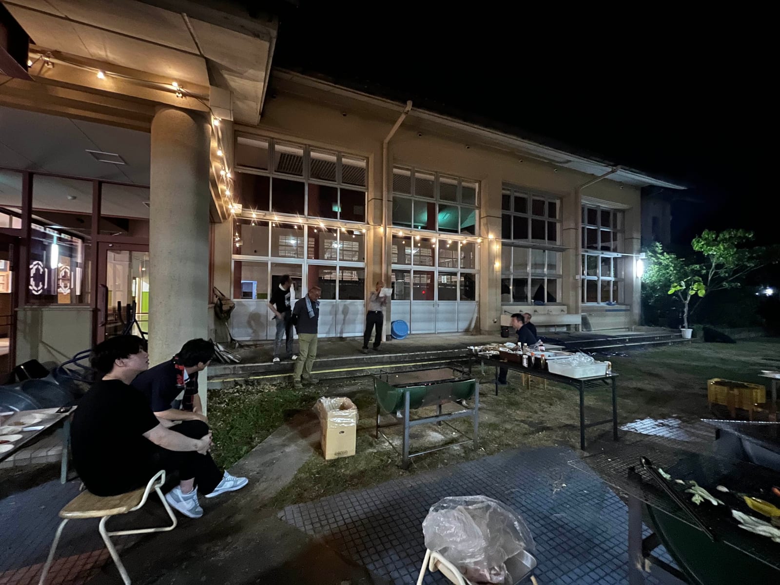 夜、灯りのついた渡り廊下の下で続くBBQ
